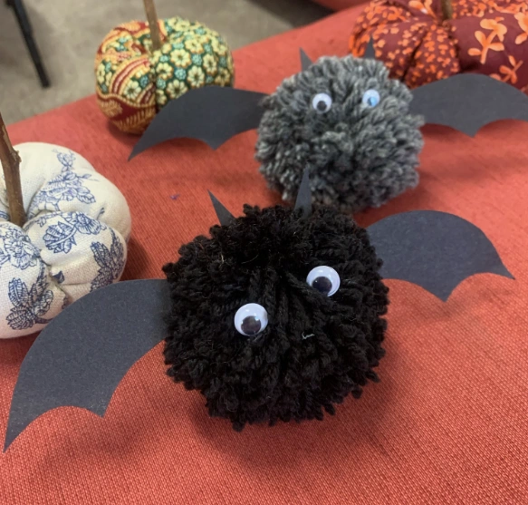 Two fluffy pom-pom bats with googly eyes and paper wings, set against a red tablecloth, surrounded by decorative pumpkins.