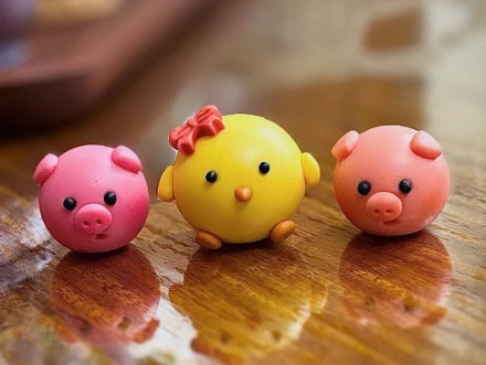 Three colorful clay figurines: a yellow chick with a bow, and two pink pigs with expressive faces, set on a wooden surface.