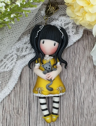 A charming clay pendant featuring a girl in a yellow dress holding a gray cat, with black hair and striped legs.