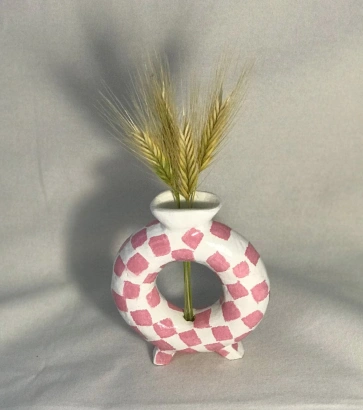 A unique clay vase with a pink checkered pattern, featuring a round base and a small opening for flowers.