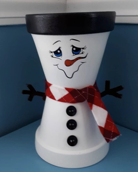 A whimsical clay pot snowman with a painted face, black top, red scarf, and stick arms, exuding a cheerful winter vibe.