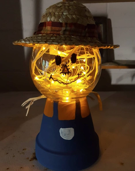 A clay pot scarecrow craft featuring a glowing bulb head with straw, wearing a straw hat, and painted blue and orange body.