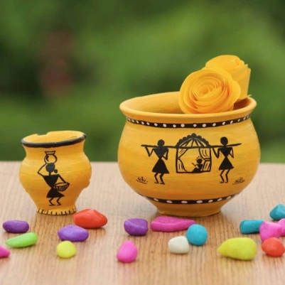 Two hand-painted clay pots in yellow, featuring black silhouettes of figures, surrounded by colorful pebbles on a wooden surface.