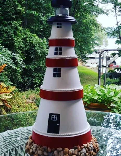 A clay pot lighthouse with red stripes and a black top, featuring small windows and a door, set outdoors among greenery.