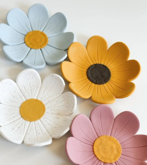 Four flower-shaped clay dishes in pastel blue, yellow, white, and pink, featuring textured petals and contrasting centers.