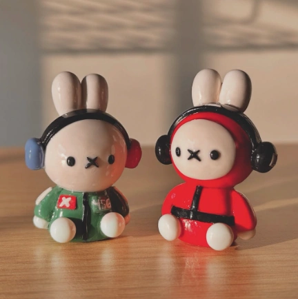 Two small clay bunny figurines, one in green and one in red, wearing headphones, sitting on a wooden surface.