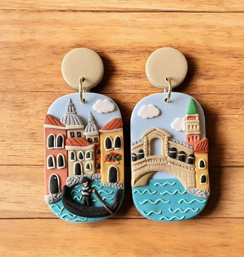 Colorful clay earrings featuring intricate Venice scenes with buildings, a gondola, and clouds, set against a blue background.
